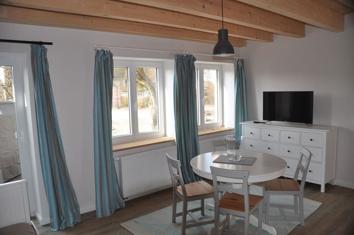 A dining area features a round table surrounded by four chairs. Large windows with light blue curtains allow natural light to fill the space. A white chest of drawers and a flat-screen TV are positioned against the wall, enhancing the room's functionality.