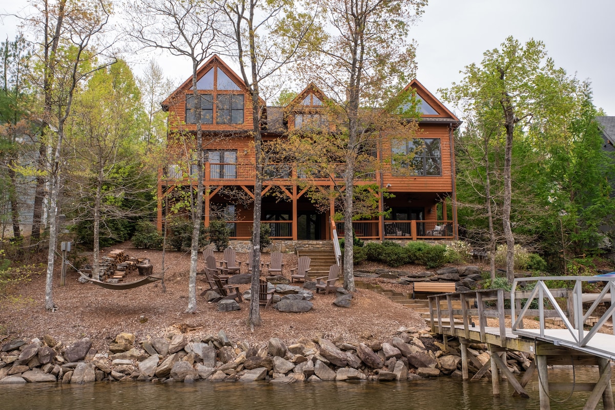 A spacious lakefront home features large windows and multiple levels, surrounded by greenery and a rocky shoreline. An outdoor seating area with chairs is present on the deck, inviting relaxation. A private boat dock extends into the lake, while trees offer a natural backdrop.