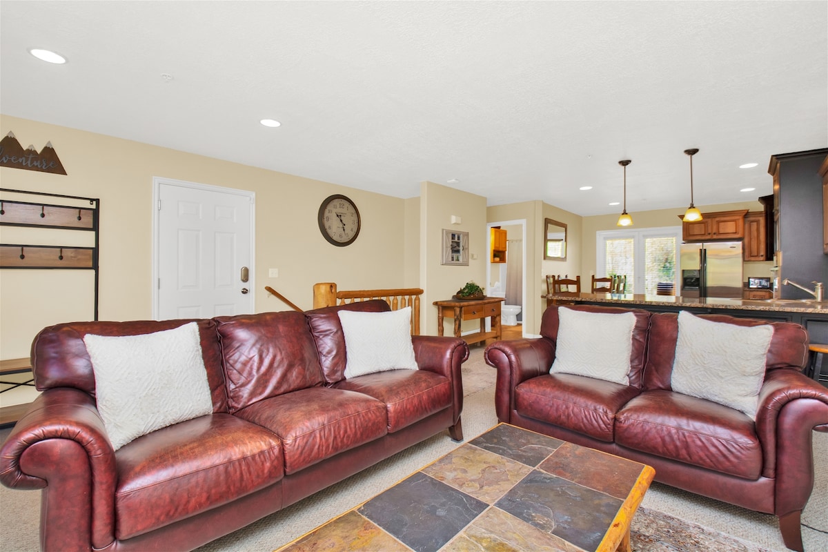 The living area features two brown leather sofas adorned with light-colored pillows, arranged around a stone coffee table. A wall clock is visible, along with wooden decor elements. Natural light streams in from the nearby windows, creating a spacious and comfortable atmosphere.