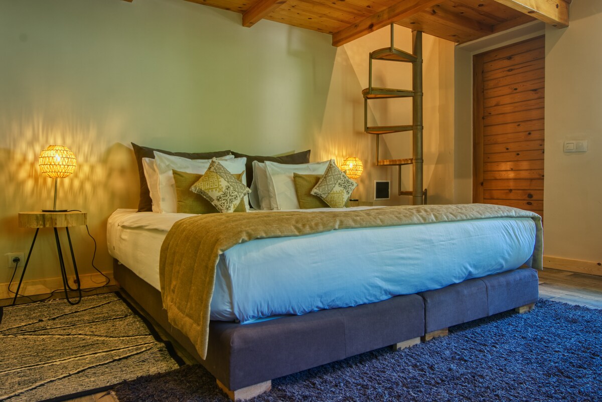 A spacious bedroom features a large bed dressed in neutral linens, complemented by decorative pillows. A cozy blanket is draped across the bed. Warm lighting is provided by two bedside lamps, and a unique spiral staircase is visible in the background.