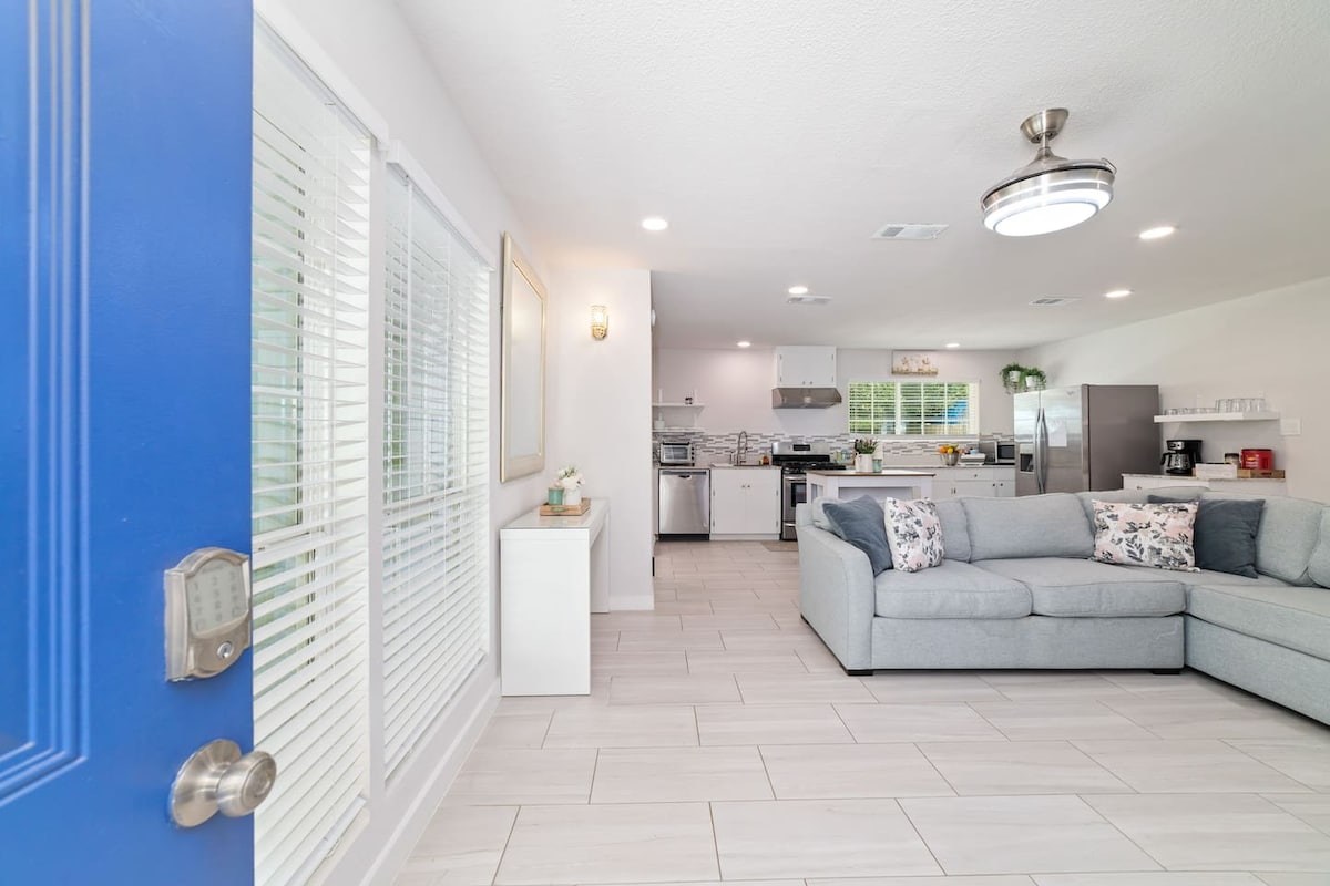 Upon entry, a spacious living area is revealed, showcasing a light grey sectional sofa adorned with floral cushions. A modern kitchen can be seen in the background, featuring stainless steel appliances and a clean, open layout. Natural light filters through the partially closed blinds.