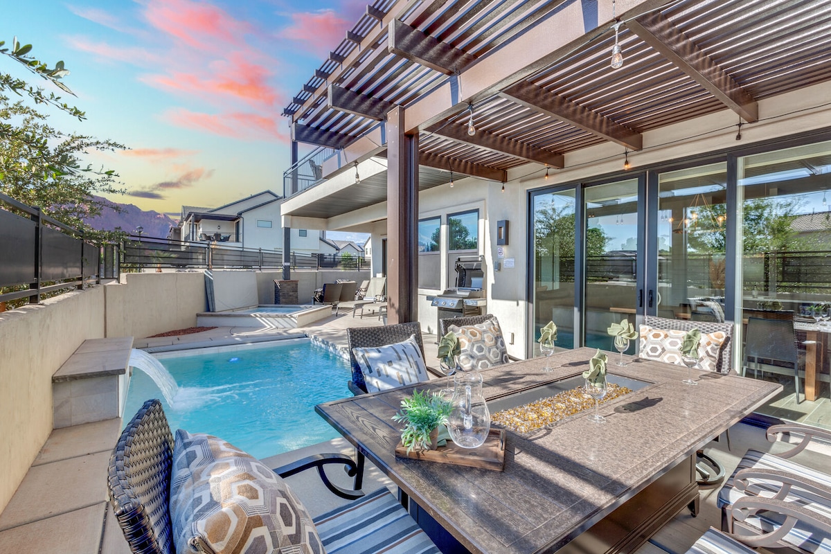 An outdoor dining area is featured beside a private pool, showcasing a large table with decorative elements. Lounge chairs are positioned around the pool, and an outdoor kitchen is visible nearby. The space is framed by modern architecture and features a backdrop of scenic hills.
