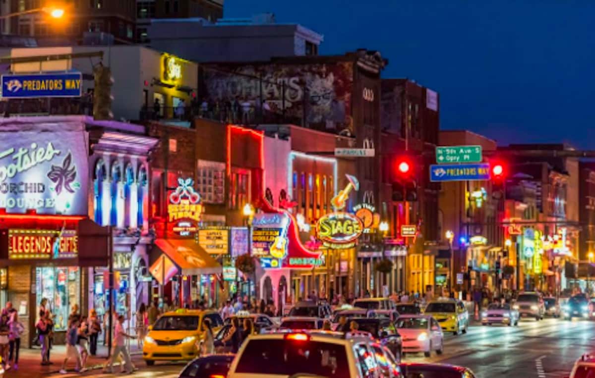 A lively street scene showcases illuminated storefronts and neon signs along a bustling avenue. Vibrant colors reflect off the vehicles parked on the road, while pedestrians enjoy the energetic atmosphere during the evening in this popular urban area.