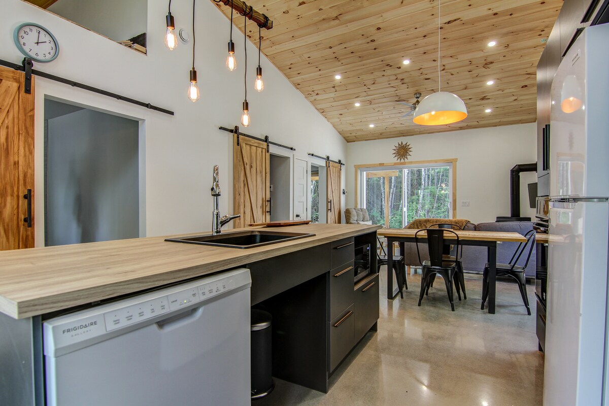 A modern kitchen features wooden cabinetry, a large island with a built-in sink, and black metal chairs around a dining table. Natural light floods the space through expansive glass doors leading to a forested area, while pendant lights illuminate the room.