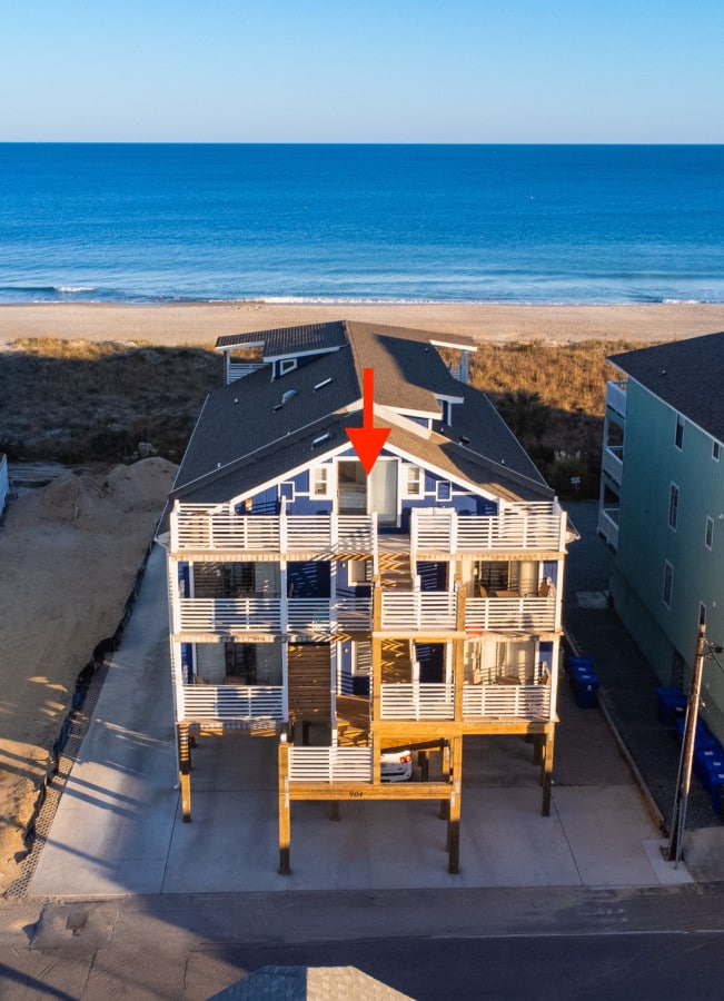 An aerial view of the beachfront condo illustrates a three-story wooden structure with multiple balconies. The expansive ocean can be seen in the background, along with a clear sky. A parking area is visible in front of the building, with several blue bins lined up along the side.
