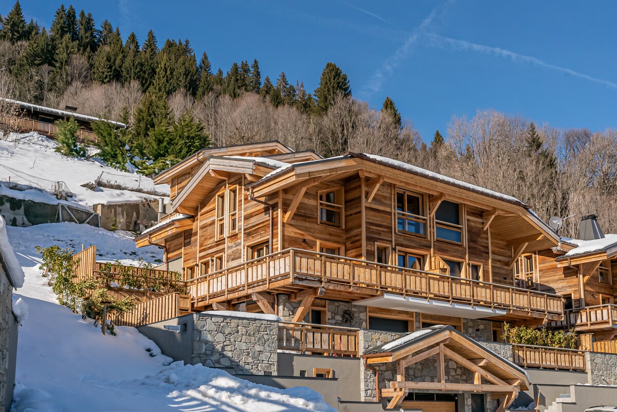 The wooden exterior of the chalet features multiple balconies and large windows, showcasing an inviting blend of modern design and natural materials. Snow covers the ground, while evergreen trees rise in the background against a clear blue sky.