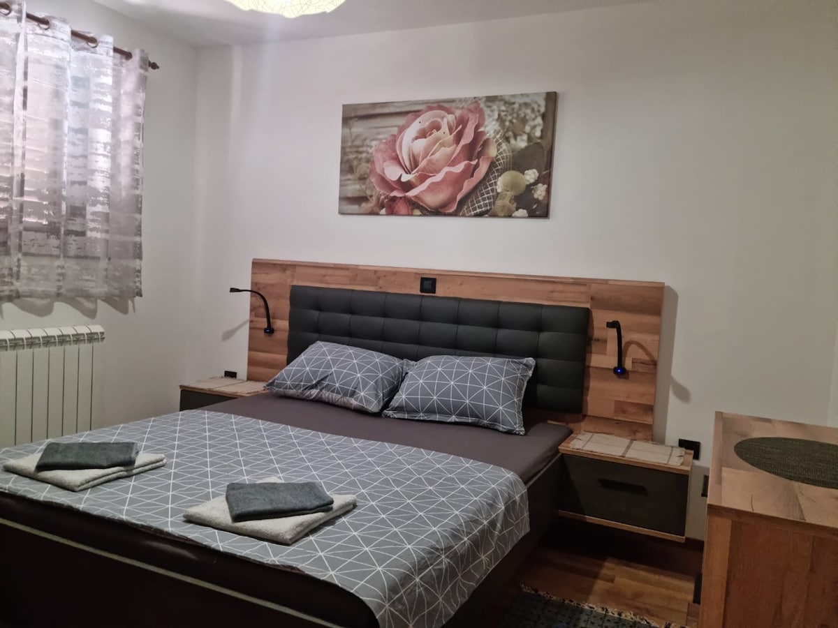 The sleeping area features a spacious bed with a modern headboard and two bedside lamps. A floral artwork adorns the wall above the bed, and neatly arranged towels are placed at the foot of the bed. Natural light filters through the sheer curtains.