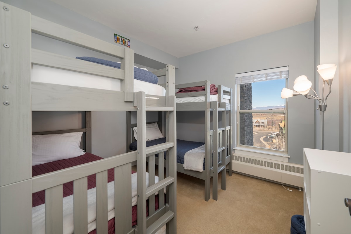 Спалня с два комплекта дървени единични легла над двуетажни легла и прозорец с изглед към планината.