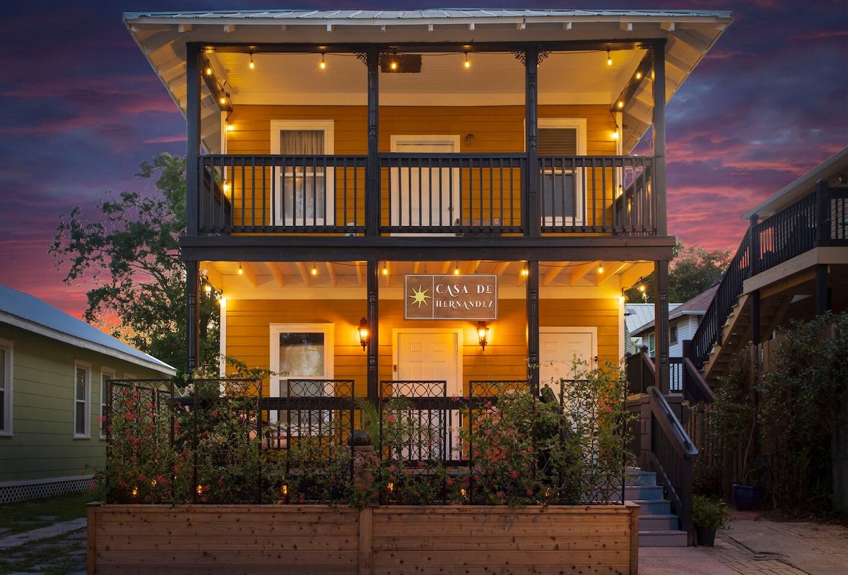The two-story Casa de Hernandez exhibits vibrant yellow exterior walls and black trim. The property features a welcoming front porch with decorative railings and layered lighting. The flowerbed in front is filled with greenery, enhancing the inviting presence of the house during twilight.