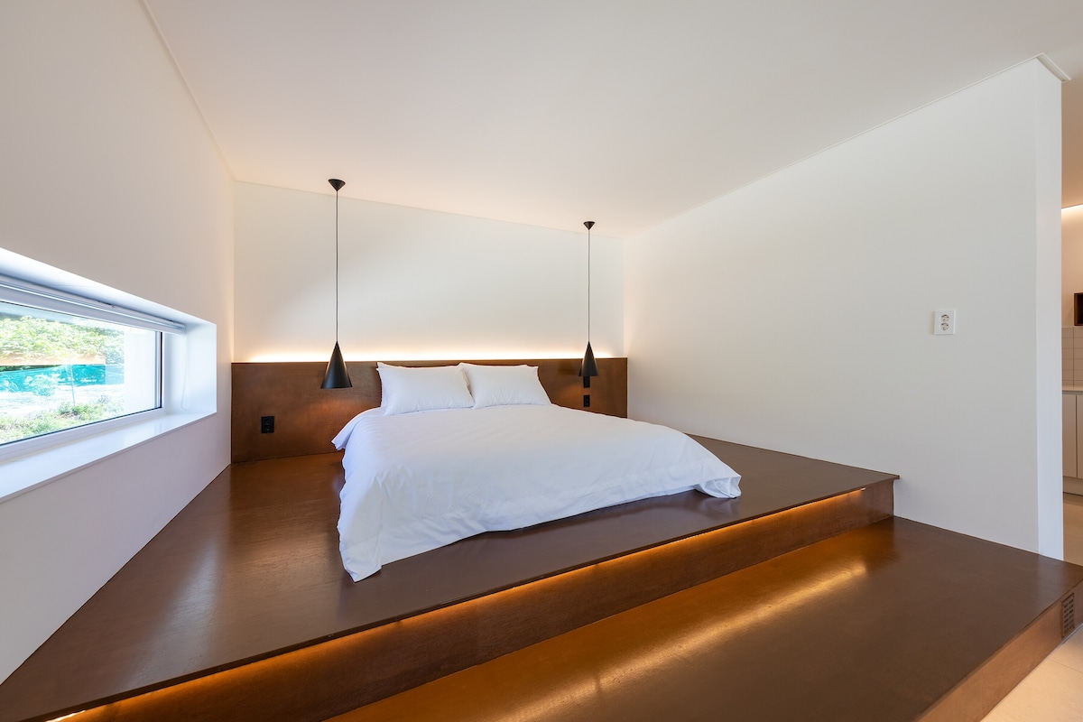 A spacious sleeping area is set on an elevated platform, featuring a large mattress with crisp white linens. Two hanging pendant lights provide ambient lighting, and a recessed window allows natural light to brighten the room.