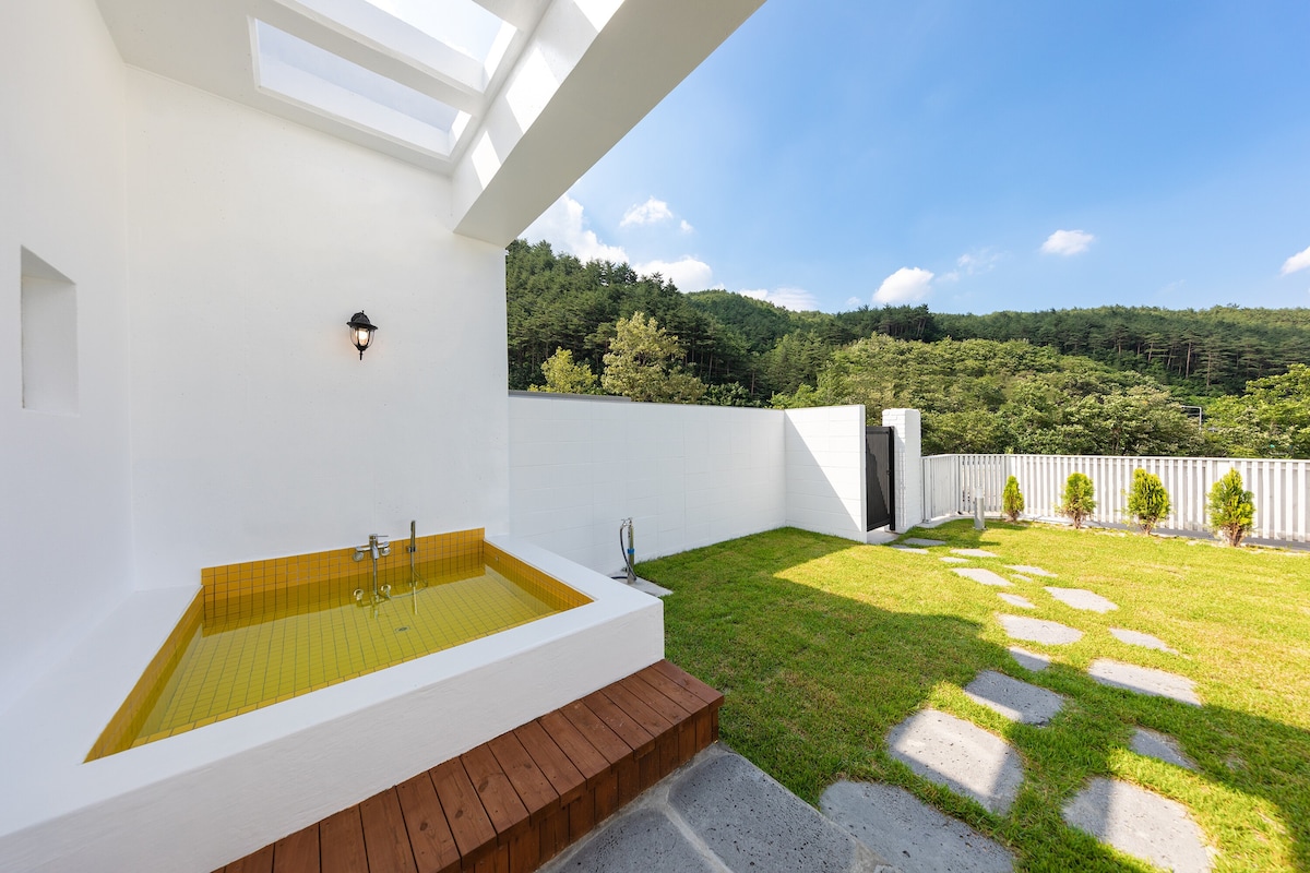 An outdoor space features a wooden deck leading to a yellow-tiled bath and a spacious grass area surrounded by stone pathways. Lush greenery and trees provide a serene backdrop, while the sky above is bright with a few clouds.