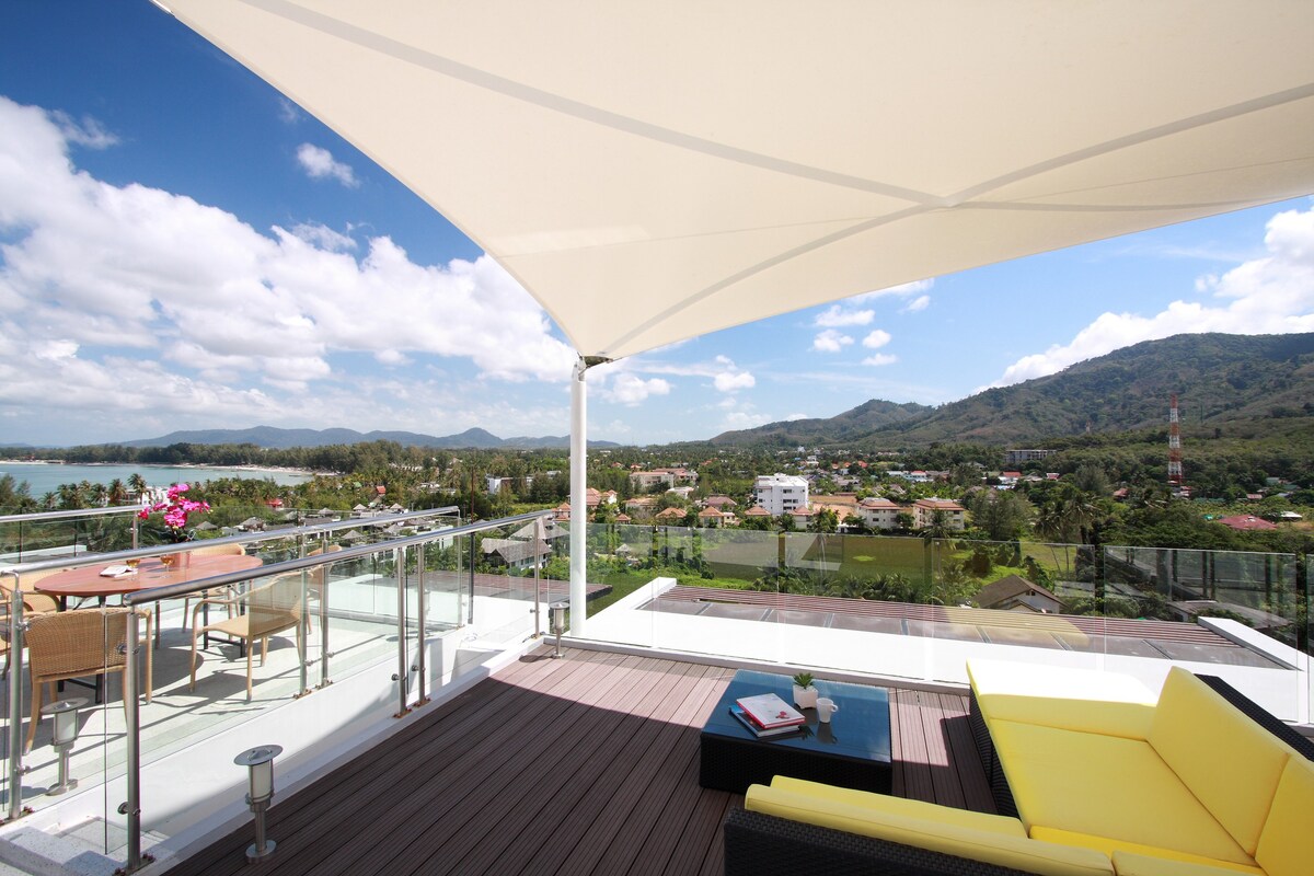 An expansive rooftop terrace features comfortable outdoor seating and a dining area. A large canopy provides shade over the space. Panoramic views of lush mountains and the coastline can be seen in the distance, with scattered village homes nestled in the landscape.