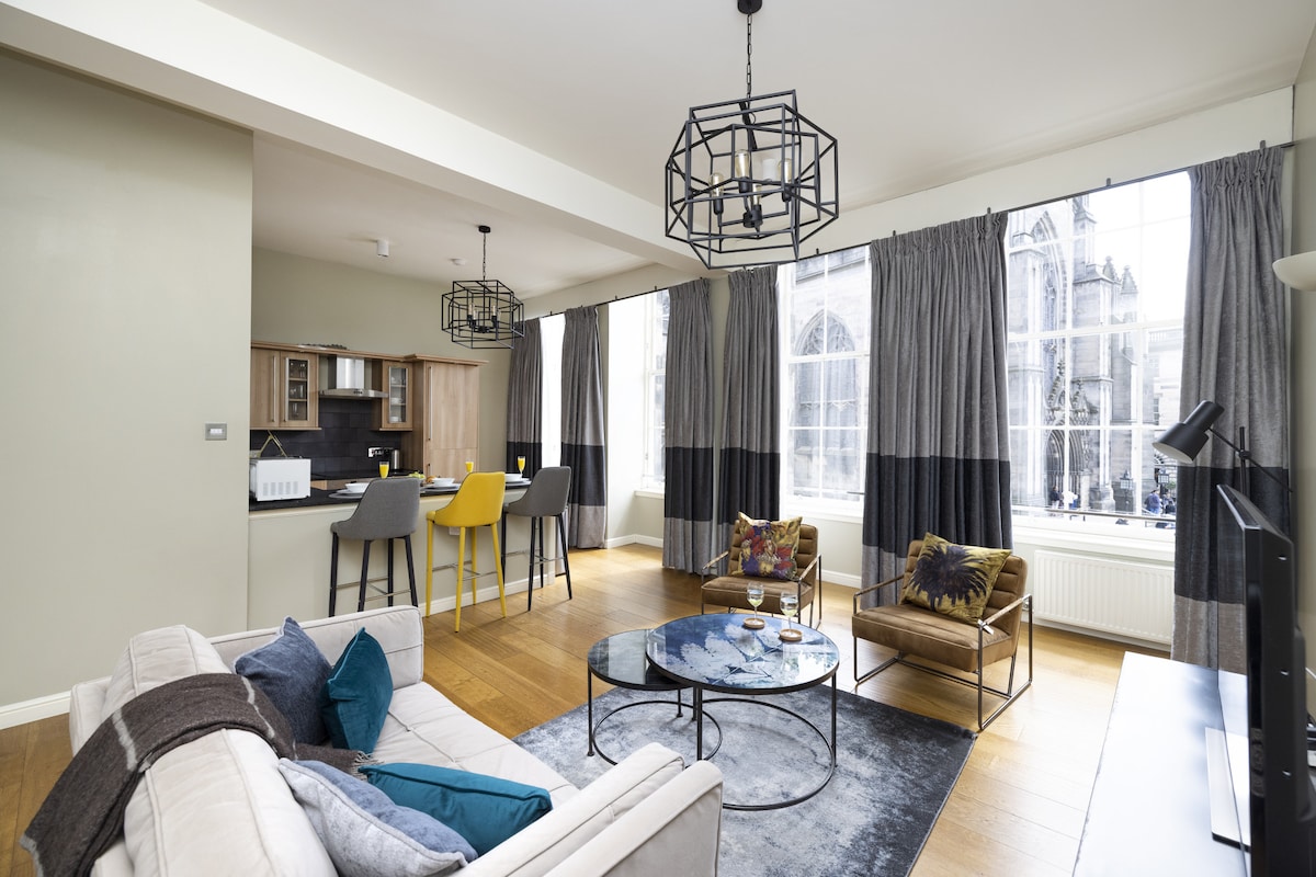 The spacious living area is illuminated by large windows, allowing natural light to fill the room. Comfortable seating is arranged around a circular coffee table, with decorative cushions adding color. The open kitchen can be seen in the background, featuring modern appliances and bar seating.