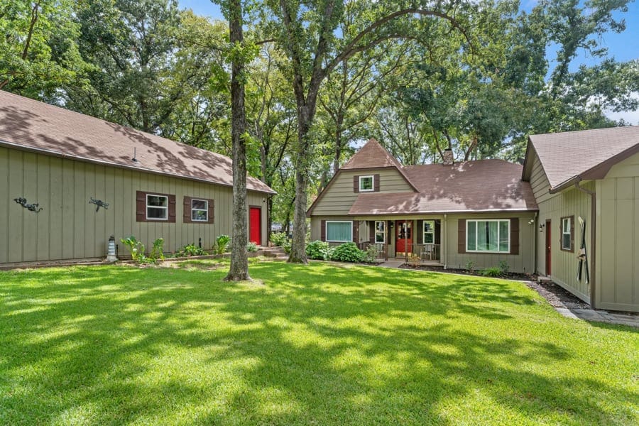 The exterior features a spacious lawn surrounded by mature trees, offering a serene atmosphere. Two buildings are visible: the main house with a peaked roof and a detached guest house with a bold red door. Greenery enhances the inviting outdoor space.
