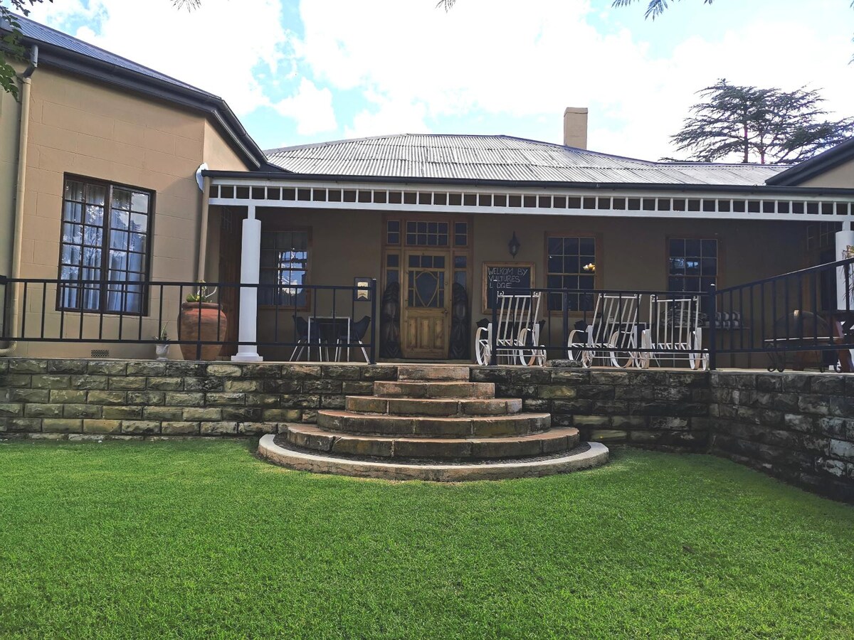 A welcoming entrance features a stone staircase leading to a covered porch, adorned with white rocking chairs. The exterior showcases light-colored walls and contrasting dark shutters, surrounded by a well-manicured lawn and trees that provide a natural backdrop.
