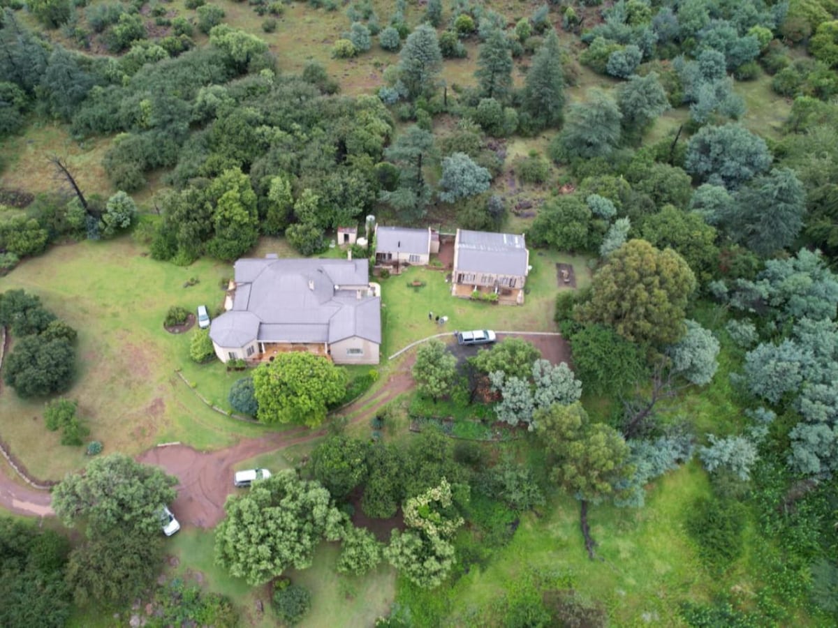 Aerial view of a self-catering cottage surrounded by lush greenery, featuring multiple buildings set within a natural landscape. Paths meander through the area, connecting the structures and inviting exploration of the scenic surroundings.