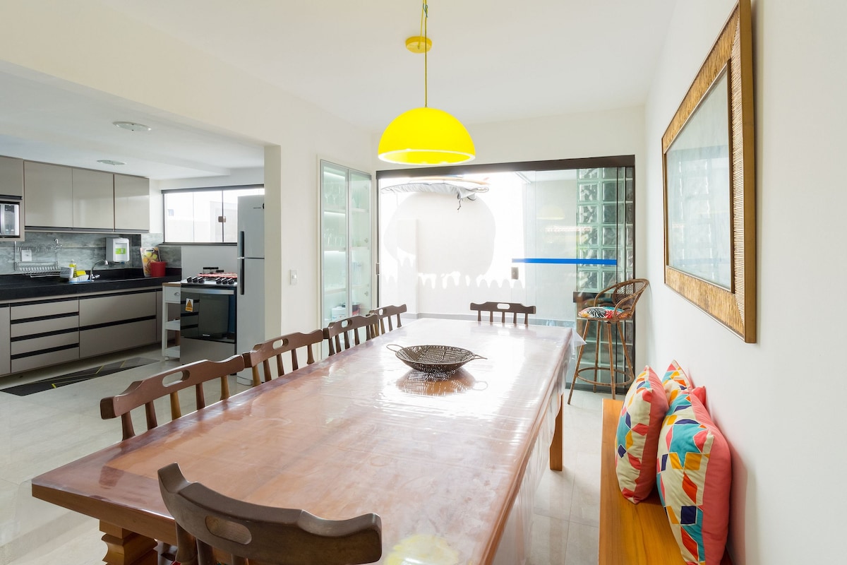 A dining area features a long wooden table surrounded by eight chairs. A vibrant pendant light hangs overhead, illuminating the space. Colorful cushions are placed along a nearby bench, with a decorative bowl positioned at the center of the table. Views of an outdoor area can be seen through the glass doors.