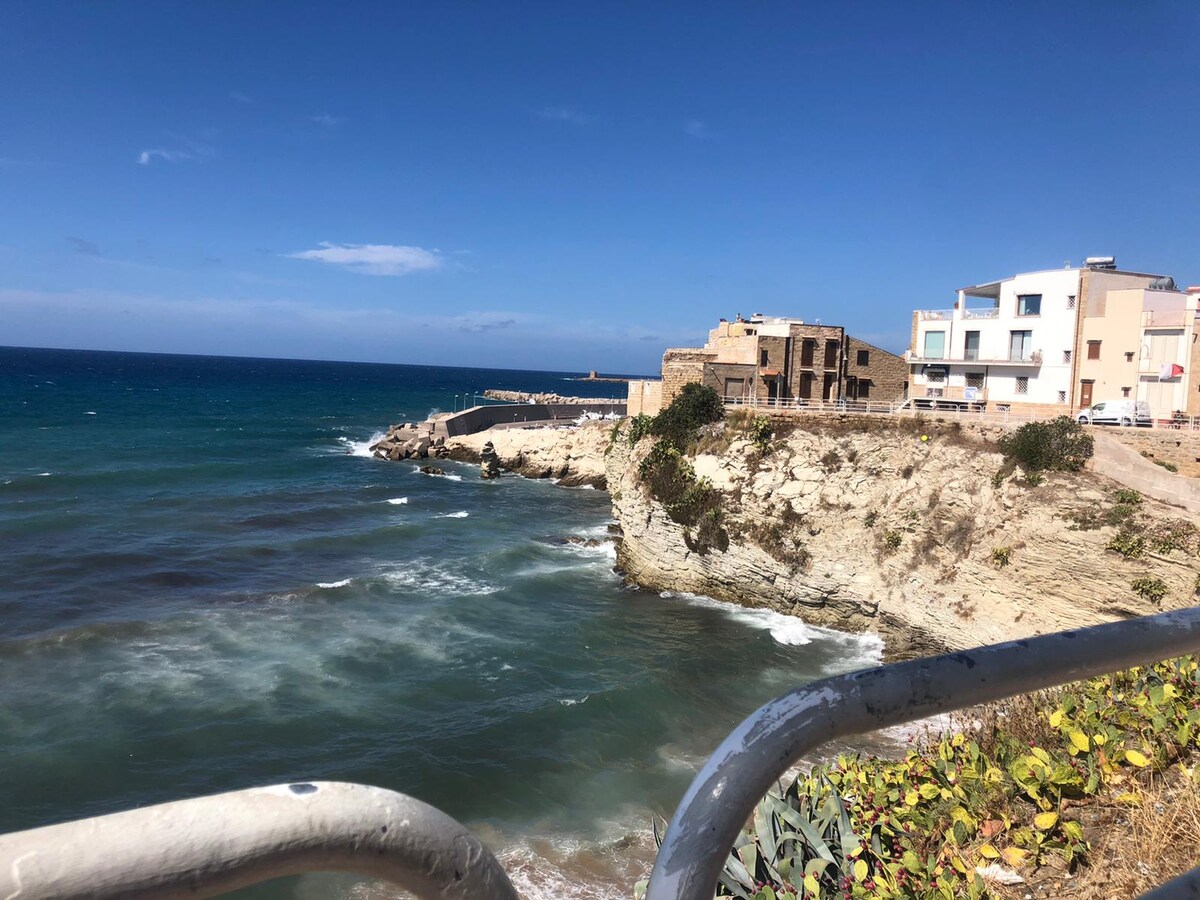 The image captures a coastal view featuring a rocky shoreline meeting the sea, with waves gently lapping against the rocks. Two distinct buildings are positioned alongside the water, highlighting the proximity of the property to the scenic coastline.