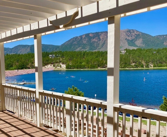 A spacious outdoor viewing deck features a white railing overlooking a serene lake surrounded by lush greenery and mountains. Bright blue skies reflect on the water, while kayakers can be seen enjoying the lake's tranquility.
