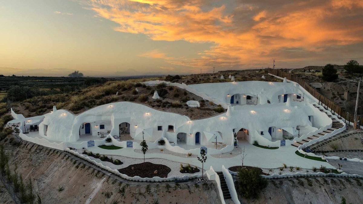 A series of uniquely designed cave accommodations is showcased against a vibrant sunset. The white exteriors blend harmoniously with the natural surroundings, featuring blue doors and windows. Paths lead to each entrance, enhancing the tranquil setting among hills and greenery.