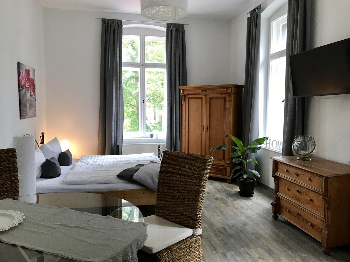 A bright room features a large bed adorned with light bedding, accompanied by decorative pillows. Natural light floods through tall windows, with elegant grey curtains framing them. A wooden wardrobe and a vintage sideboard provide storage, while a small dining area includes woven chairs and a table.
