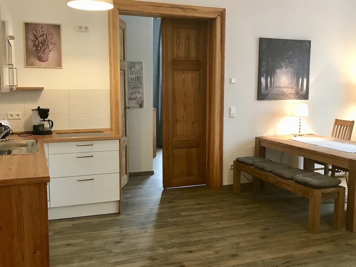 The open-plan living kitchen area features wooden cabinetry and a kitchenette equipped with a coffee maker. A dining table with bench seating is present, illuminated by soft light from a nearby lamp. Artwork depicting a forest scene can be seen on the wall.