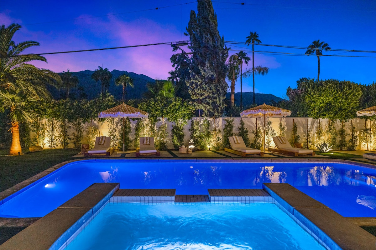 The inviting pool area is illuminated under a twilight sky, showcasing vibrant blue waters. Lounge chairs are arranged beside the pool, with umbrellas providing shade. Lush greenery lines the space, enhancing the sense of tranquility, while majestic palm trees rise in the background.