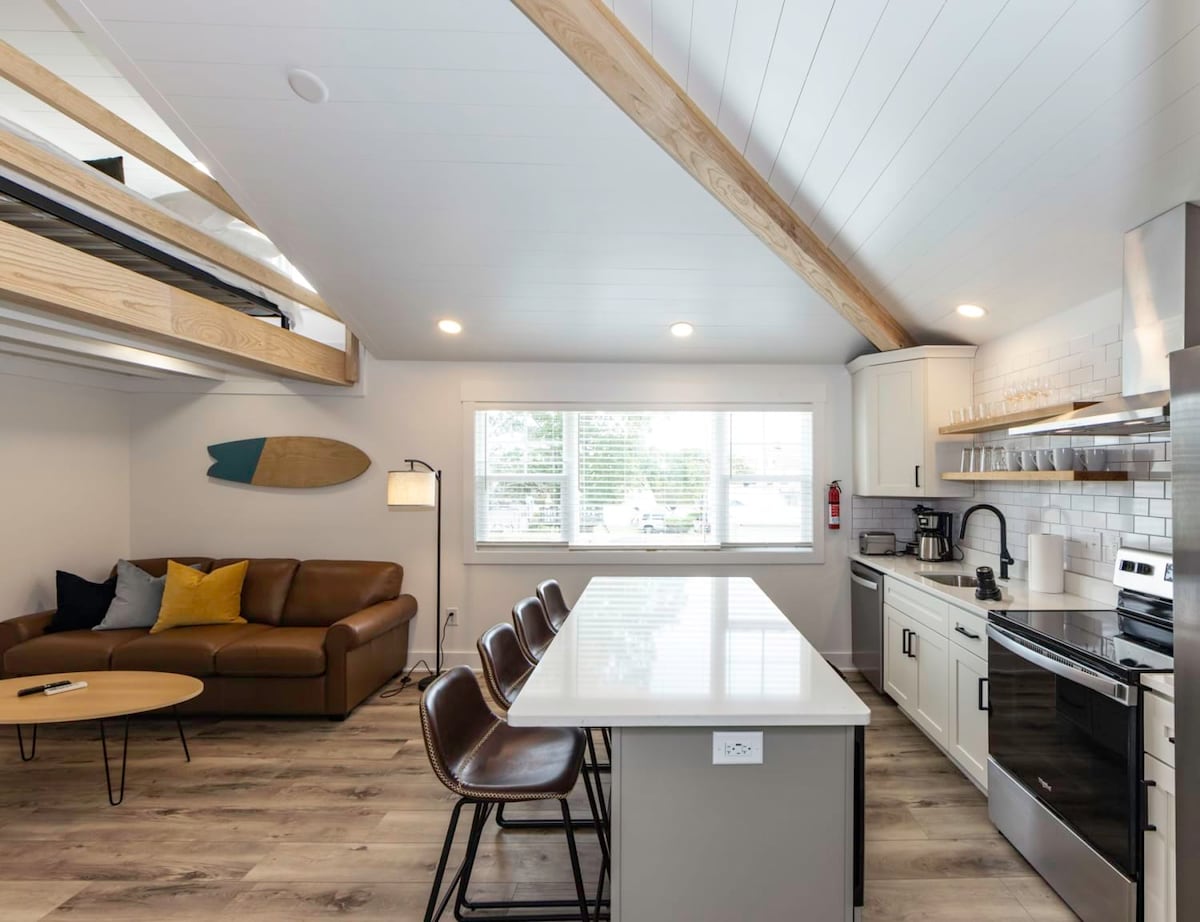 A modern interior space features a well-equipped kitchen with stainless steel appliances and a bright island bar. A comfortable brown sofa is positioned near large windows that invite natural light. A lofted sleeping area with a minimalist design can be seen above, alongside decorative surfboards.