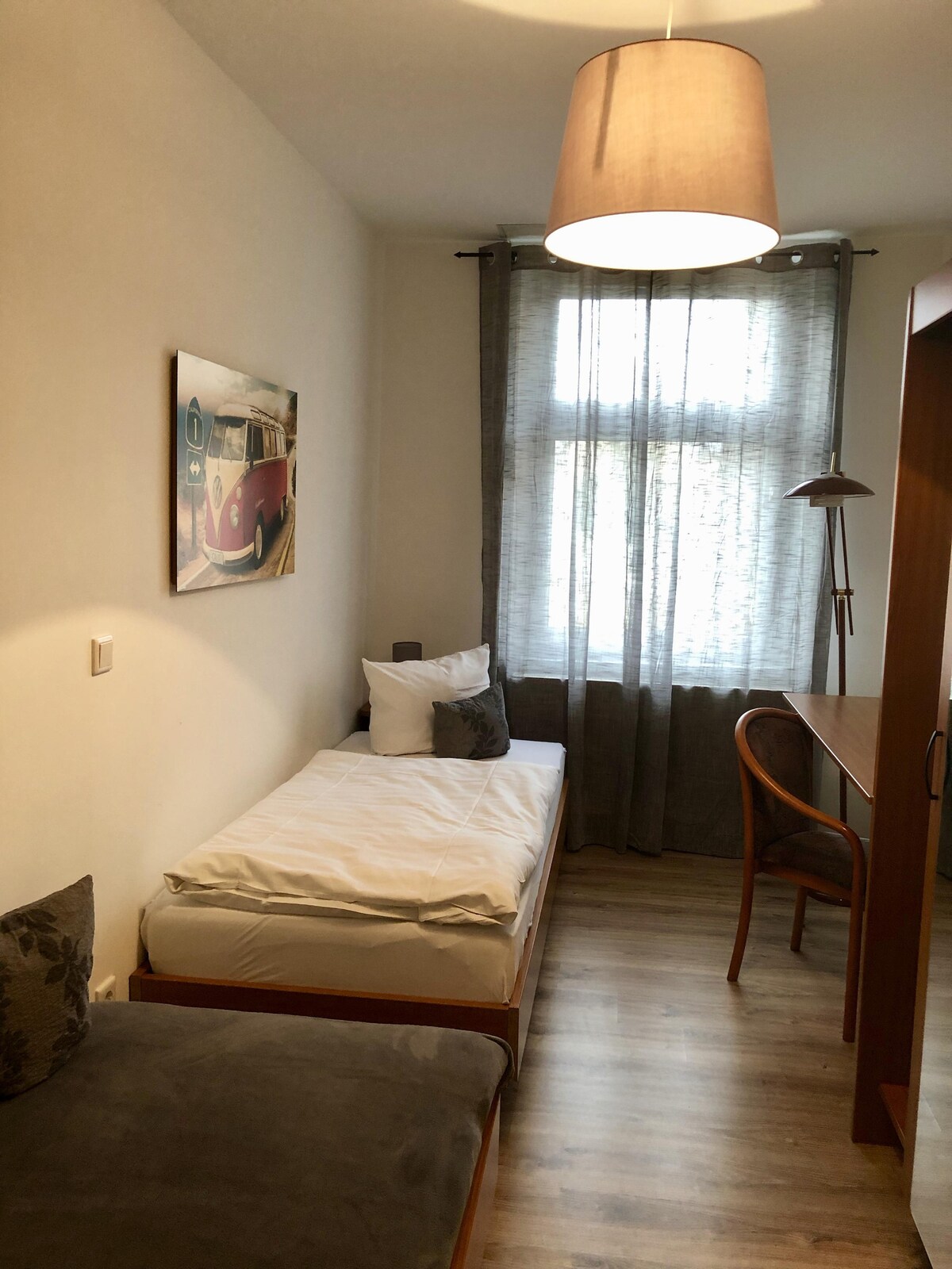 A cozy bedroom features a single bed against a light wall. A decorative photograph of a vintage car hangs above the bed. A wooden desk and chair are positioned nearby, and soft natural light filters through sheer curtains in front of a window.