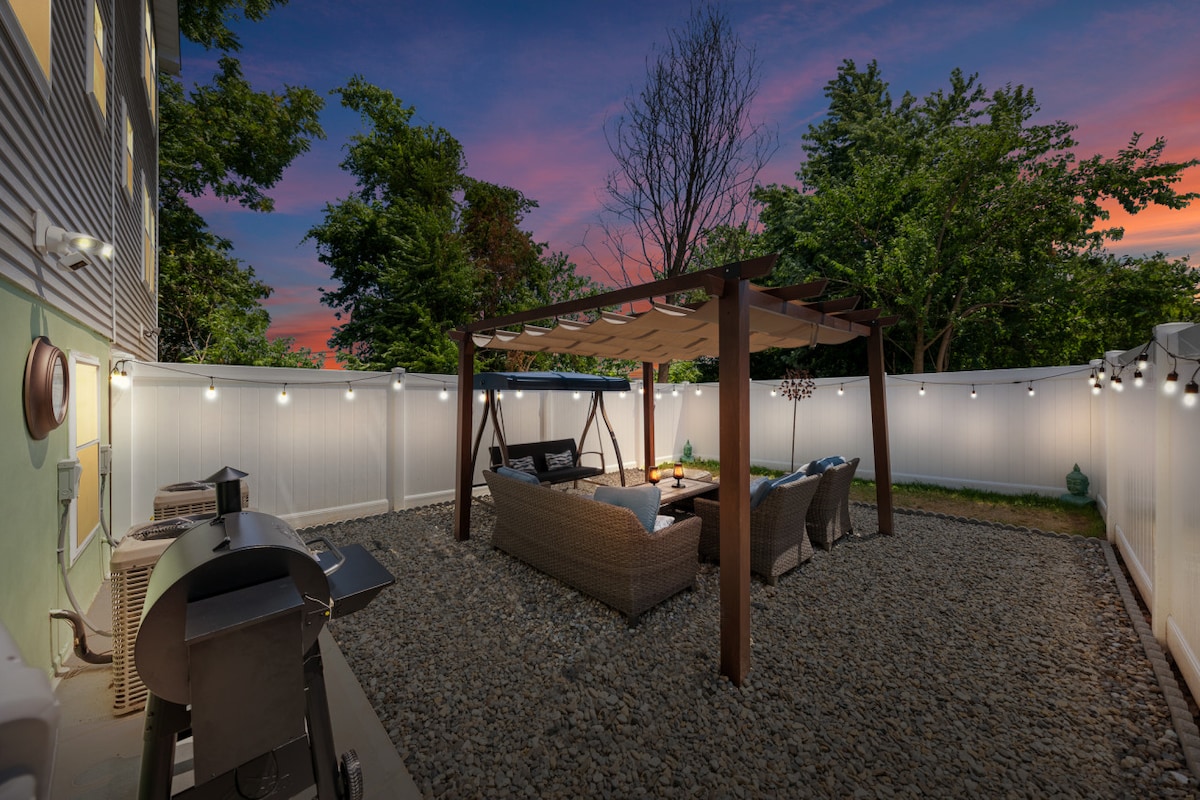 An outdoor space features a wooden pergola with comfortable seating and a coffee table, surrounded by lush greenery. Soft lighting enhances the ambiance, while a grill adds functionality for outdoor cooking. Gravel covers the ground, offering a natural, inviting area for relaxation.
