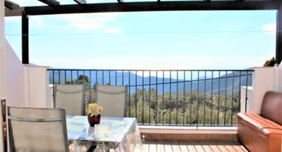 The terrace is furnished with a glass-top table and two chairs, offering a scenic view of the mountains and surrounding greenery. A comfortable sofa complements the relaxing outdoor space, ideal for enjoying the landscape and fresh air.