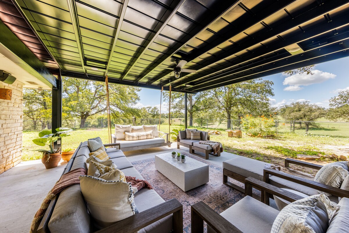 An outdoor patio space features a large covered area with comfortable seating arrangements, including a sofa and chairs. A coffee table sits in the center, surrounded by greenery and distant trees. The area is well-lit, inviting relaxation and social gatherings.