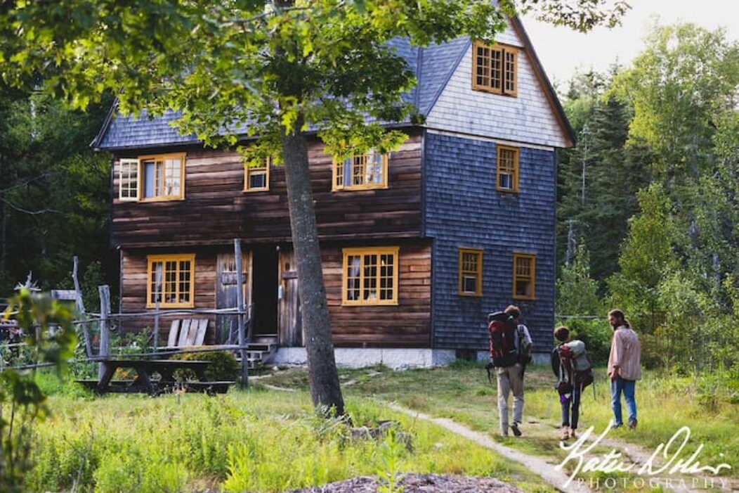 A handmade 17th Century building stands among lush greenery, featuring wooden siding and large, framed windows. Guests are seen approaching the entrance, carrying backpacks, along a grassy path that invites exploration of the surrounding nature.