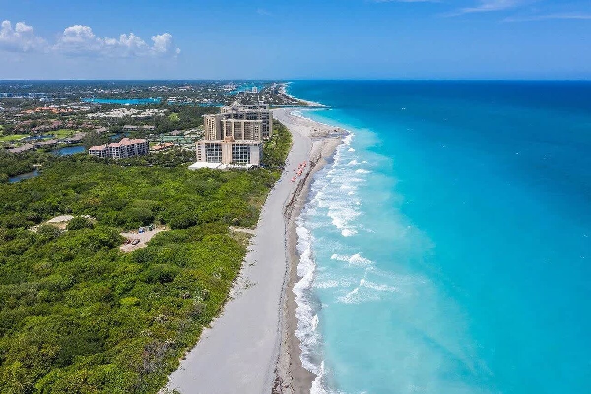 An aerial view reveals a stunning stretch of sandy beach meeting bright turquoise waters. Lush greenery outlines the beach, with a glimpse of nearby buildings along the coastline. The scene showcases a peaceful setting, inviting visitors to explore the natural beauty of the area.