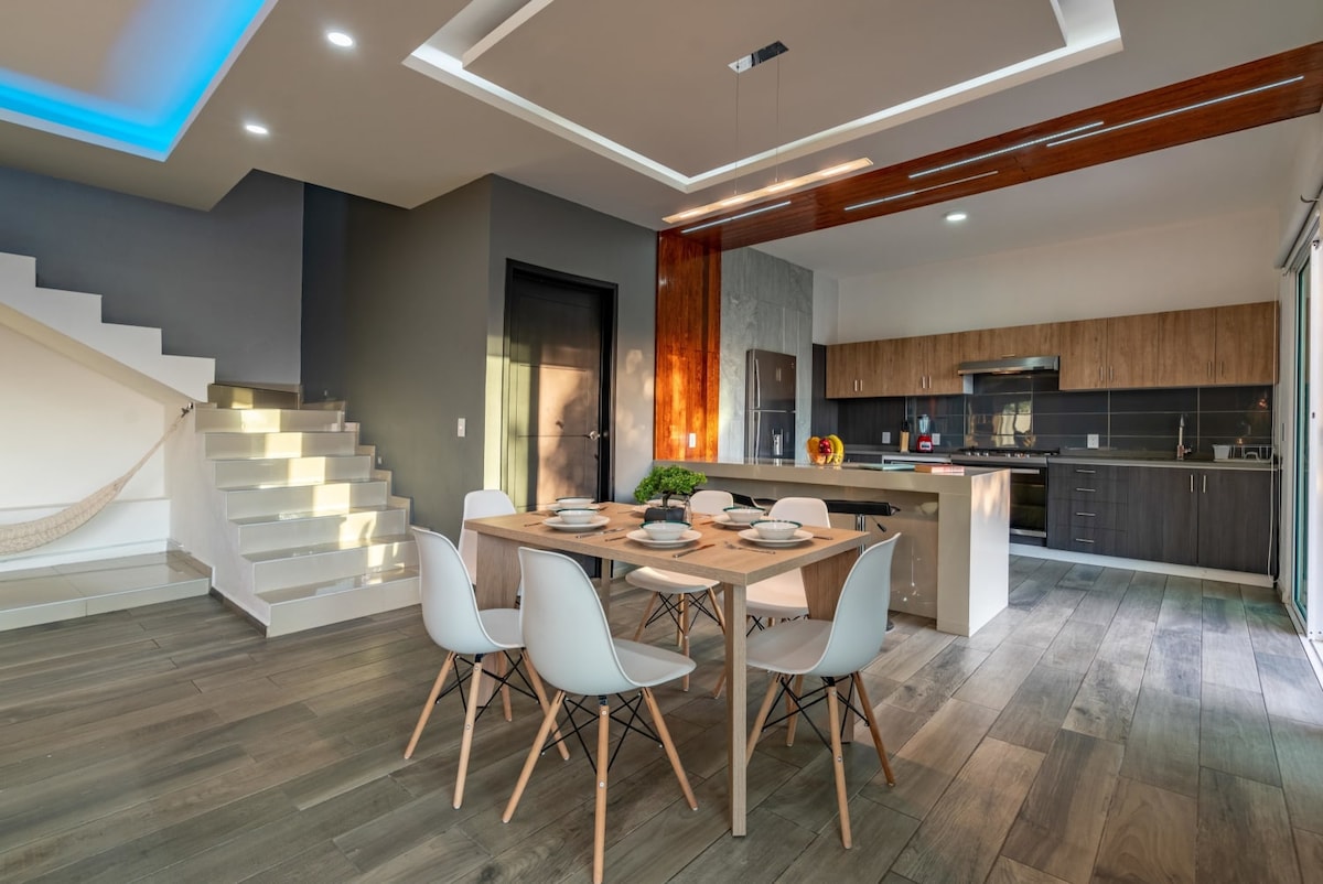 A modern dining area features a light wooden table set for four, surrounded by white chairs. Natural light flows through large sliding doors leading to an outdoor space. The kitchen showcases dark cabinetry, stainless steel appliances, and a minimalistic aesthetic.