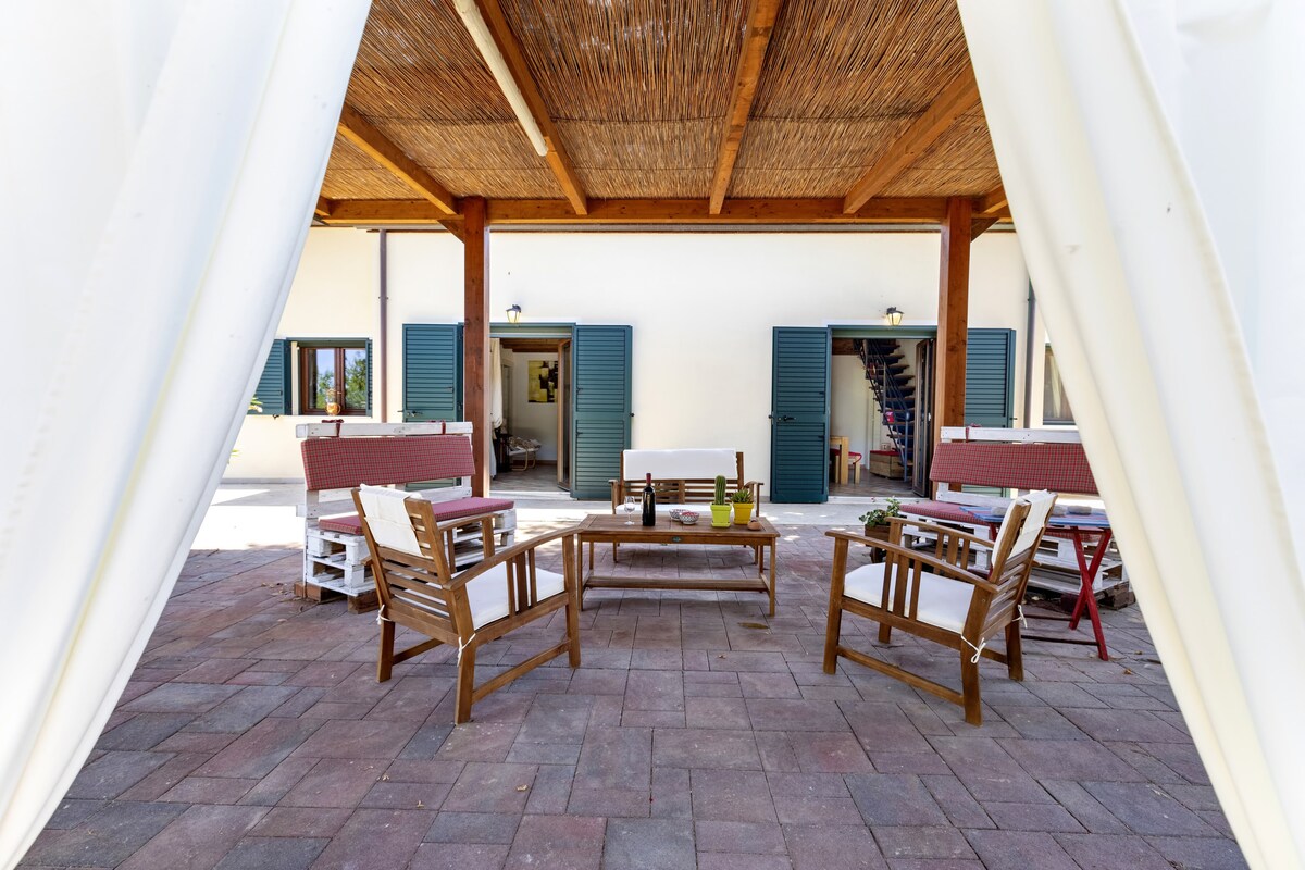 A covered terrace features wooden furniture, including chairs and a table, set on a shaded patio. The space is framed by white curtains, creating a relaxed setting. Double doors in the background lead to additional rooms, enhancing outdoor living options.