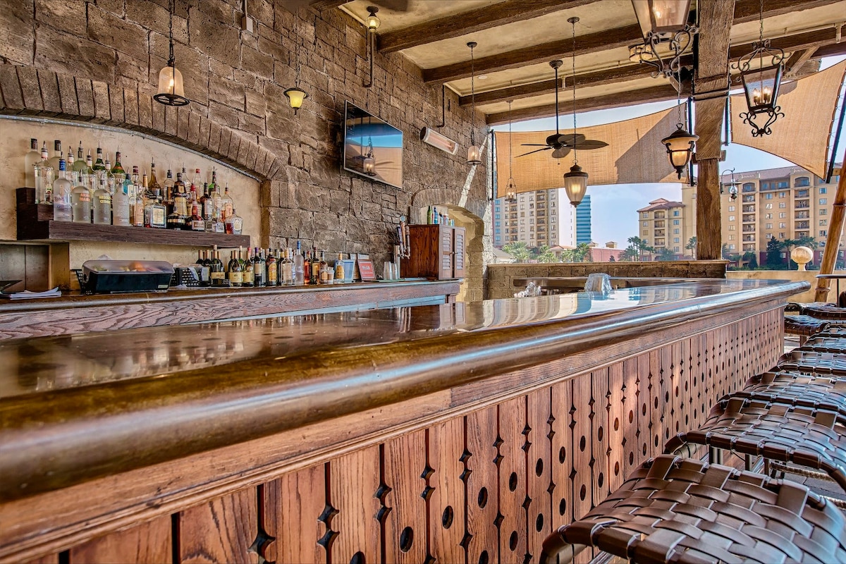 An open-air bar area features a polished wooden counter with a rich finish, accompanied by woven seating. Behind the bar, an extensive selection of bottles is displayed on shelves. Soft lighting and decorative lanterns enhance the inviting atmosphere while views of nearby buildings can be seen in the background.