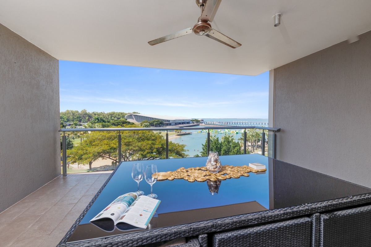 An outdoor balcony area features a glass-top dining table surrounded by woven chairs. A decorative centerpiece rests on the table along with an opened magazine. Expansive views of Darwin Harbour and the surrounding landscape are visible in the background.