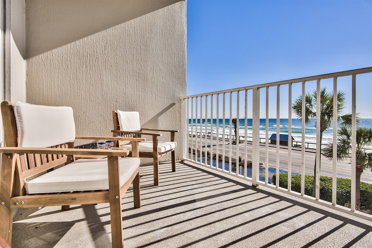A spacious balcony is furnished with two wooden chairs, providing a perfect spot for relaxation. The railing offers views of the sandy beach and the Gulf, with gentle waves visible below. Bright blue skies complement the coastal scenery, creating an inviting outdoor space.