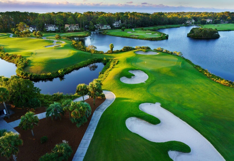 The image captures a lush golf course featuring meticulously maintained greens and sand traps. A calm body of water winds through the landscape, surrounded by trees, creating a serene environment. The scene is enhanced by the soft glow of sunlight reflecting on the water's surface.
