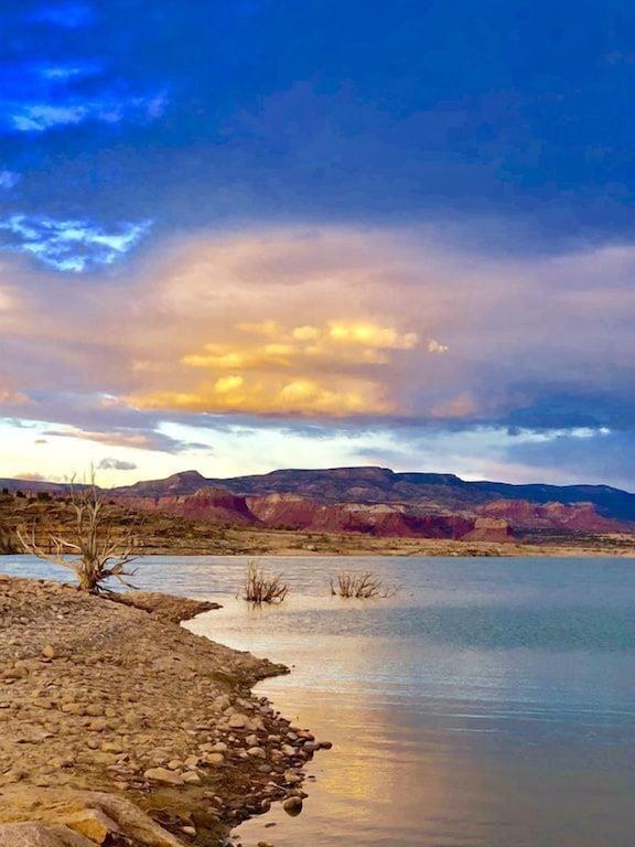An expansive view reveals a tranquil lake bordered by a gently sloping shore. The surface of the water reflects the surrounding landscape and vibrant clouds, with red rock formations in the background. A few barren trees emerge from the water, adding to the serene scene.