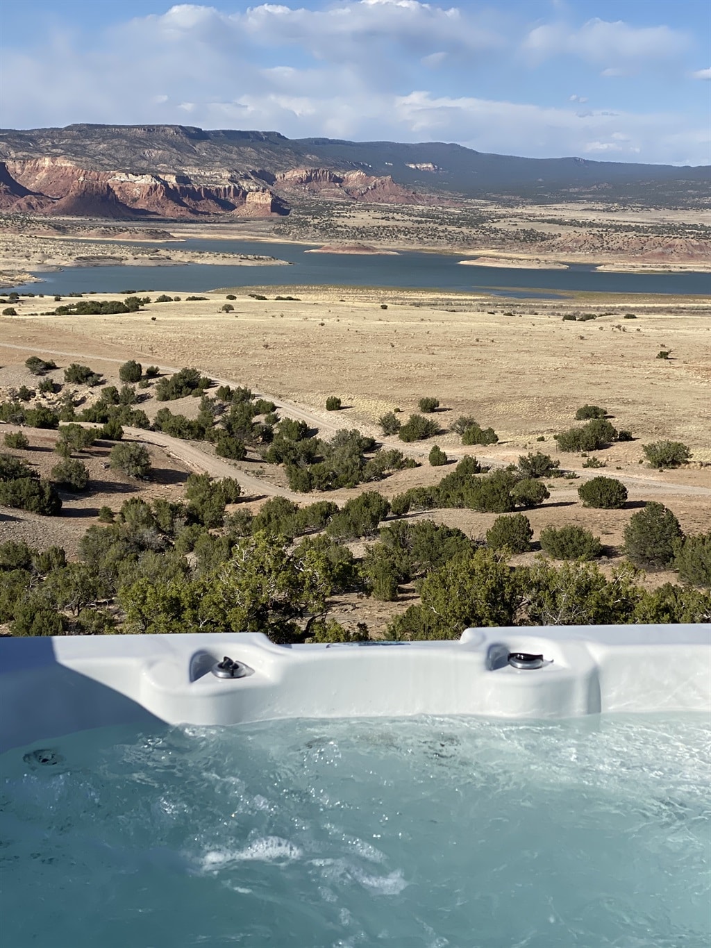 A spacious outdoor hot tub is showcased, positioned on a deck with expansive views of Abiquiu Lake and the surrounding landscape. The water is gently bubbling, with distant hills and cliffs visible in the background, creating a serene atmosphere.