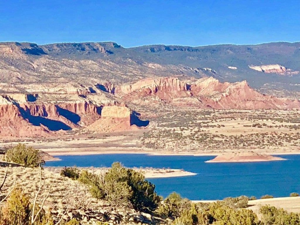 A vast landscape reveals a serene Abiquiu Lake surrounded by striking red cliffs and rugged rock formations. The water's surface glistens in the sunlight, while the backdrop features distant mountains under a clear blue sky.