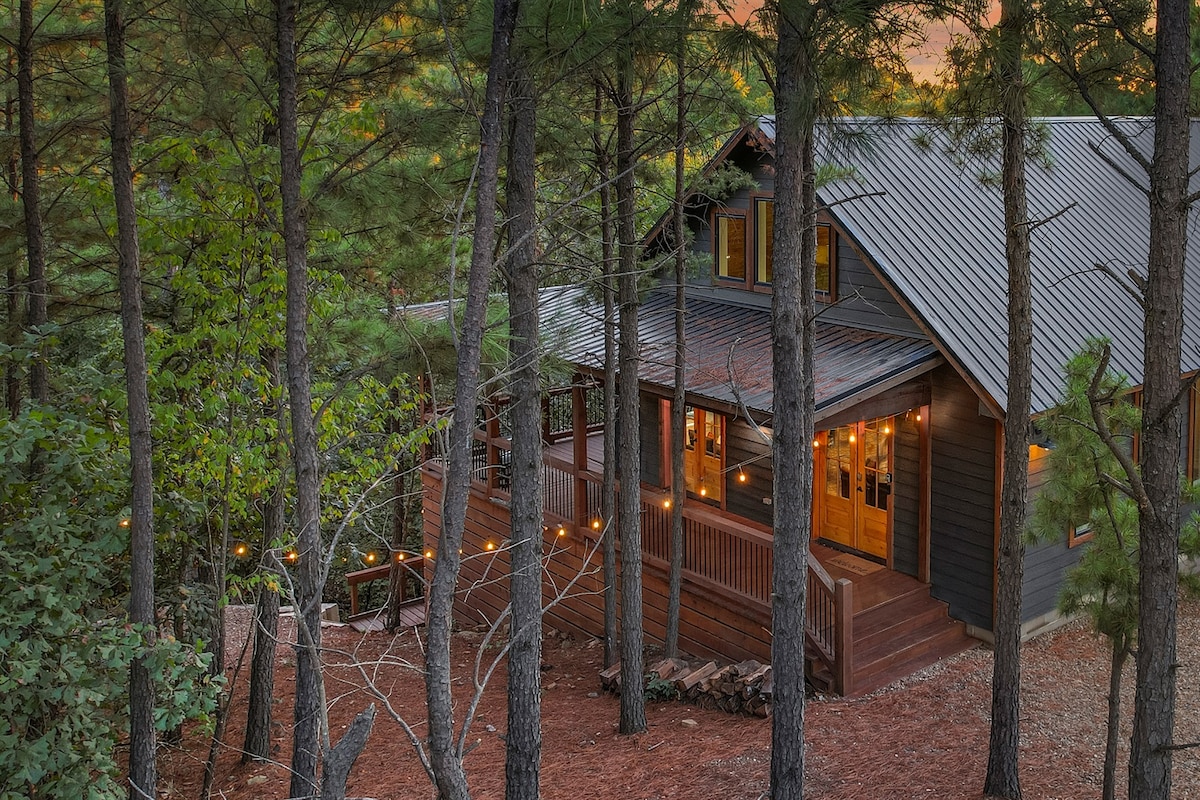 The cabin, nestled among tall pine trees, is highlighted by warm outdoor lighting. A spacious wrap-around porch can be seen, leading to the entrance. The rustic exterior features a blend of wood and metal accents, creating a harmonious look with the natural surroundings.