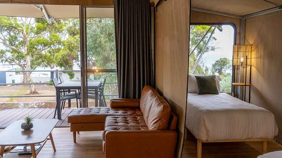 The open-plan bedroom features a queen bed and two single beds, with natural light flowing through large glass doors. A cozy seating area includes a brown leather couch and a wooden coffee table. Outside, a private deck overlooks native trees and outdoor furniture.