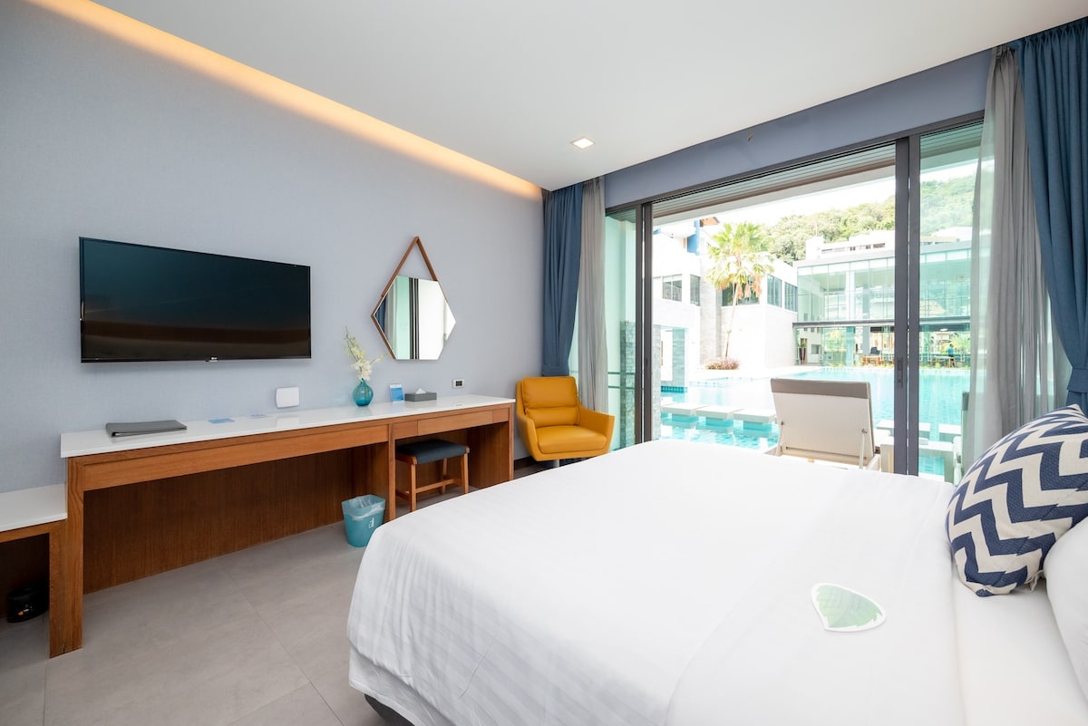 A modern room is visible, featuring a spacious bed with crisp linens and a decorative pillow. A flat-screen TV is mounted on the wall. A bright armchair sits beside a wooden desk, with large glass doors providing a view of the pool area.