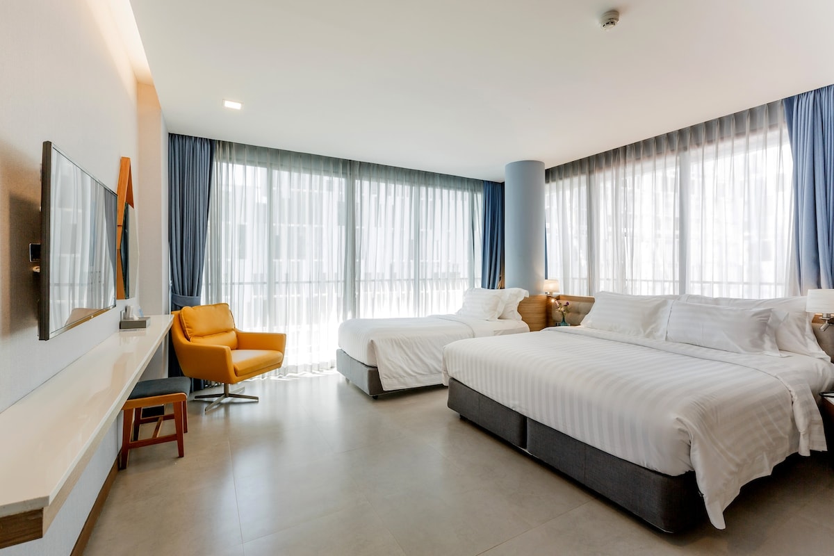 A spacious triple room features a king-size bed and a single bed, both draped in white linens. Large windows are adorned with sheer curtains, allowing soft natural light to fill the room. A comfortable chair and a flat-screen television are positioned for relaxation.