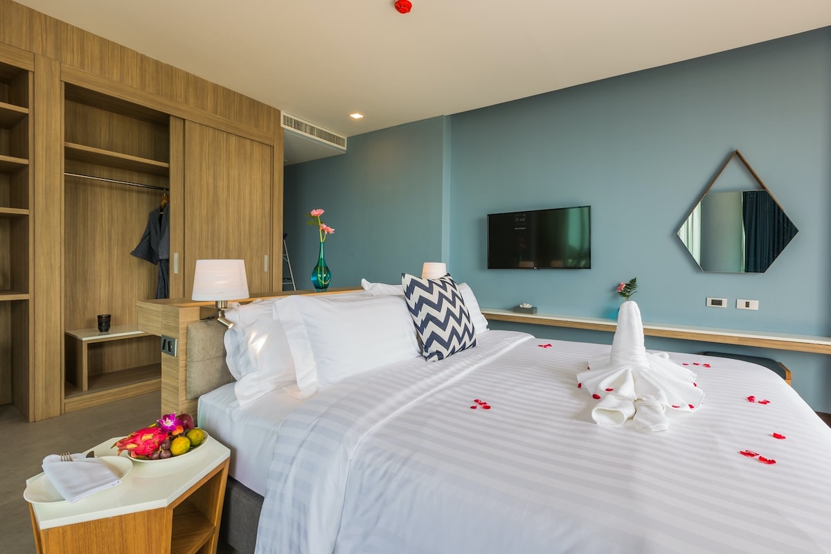 A spacious room features a king bed adorned with decorative pillows, complemented by a bedside table with a lamp. A wall-mounted television is positioned opposite the bed, and a wooden wardrobe offers ample storage. Fresh flowers and fruit are arranged on the counter, enhancing the inviting ambiance.