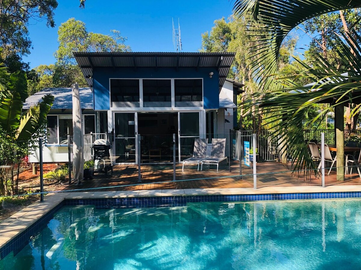 A spacious outdoor deck extends along the backside of the property, overlooking a large in-ground pool. The house, featuring large glass windows and doors, is surrounded by lush greenery, providing a serene and private atmosphere. Seating areas and a gas BBQ are visible on the deck.