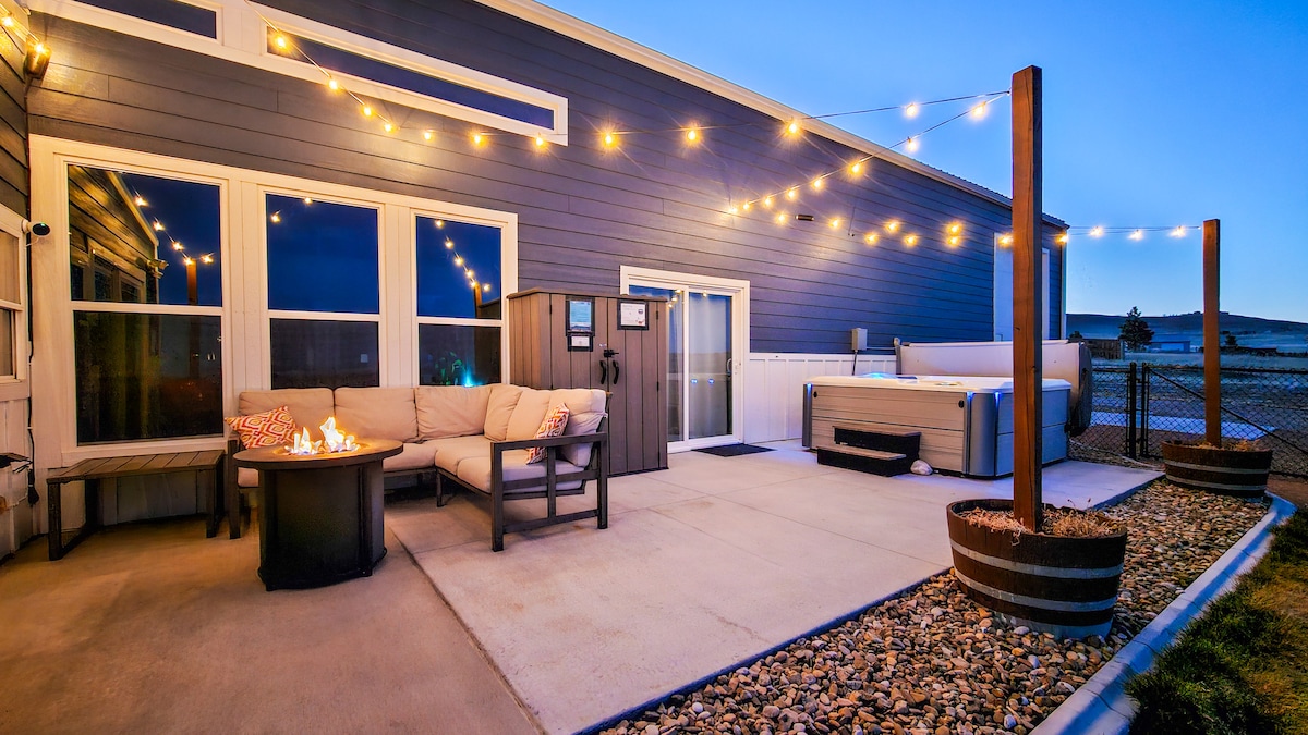 An outdoor patio area is illuminated by string lights, featuring a comfortable seating arrangement around a fire pit. A hot tub is positioned nearby, with a fenced-in backdrop and landscaped stone elements enhancing the space.